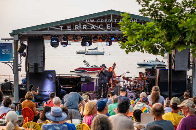 A view of a band playing on a lakefront stage as a crowd sitting on a terrace listen.