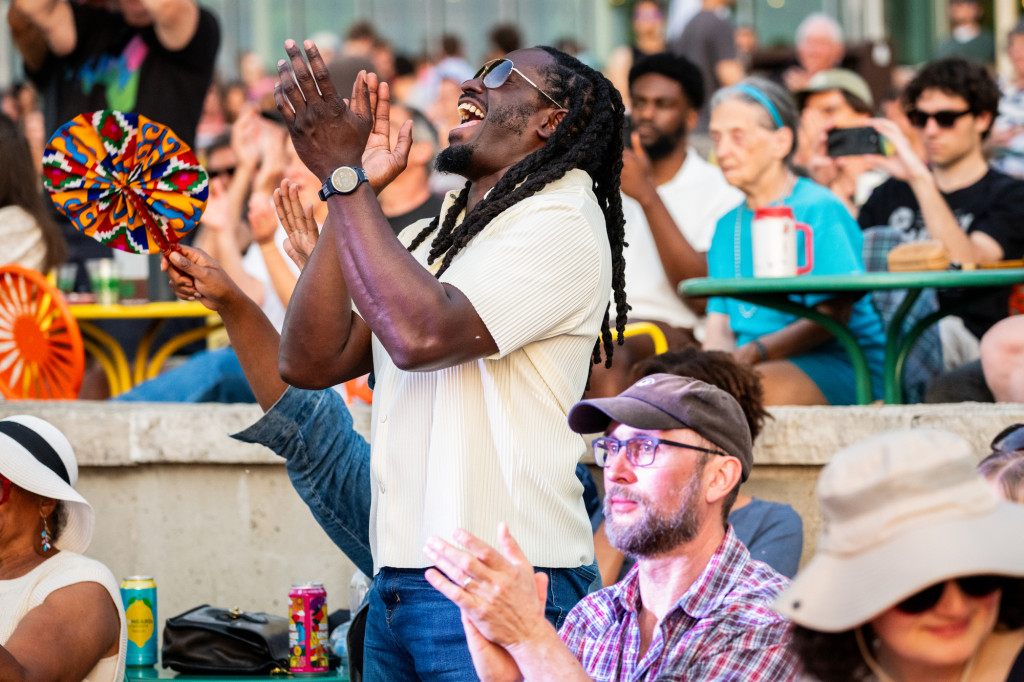 People standing and sitting on a terrace clap and cheer as a band plays.