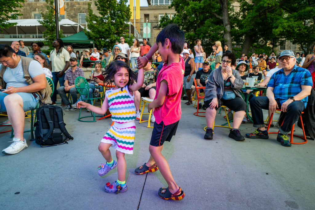 Two children dance on a terrace as people sit in chairs and listen to the music in the background.