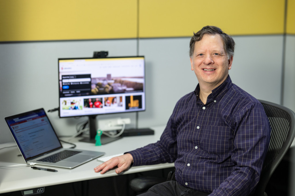 A man sits in front of a computer monitor, smiling at the camera.