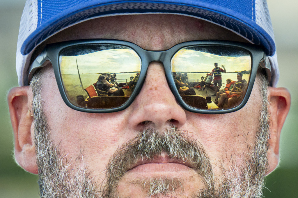 The face of a man with reflective sunglasses is seen; a lake scene is reflected in the sunglasses.