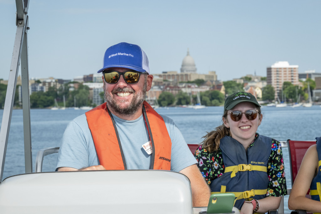 A man smiles as he steers a boat on a lake; a woman smiles in the background.