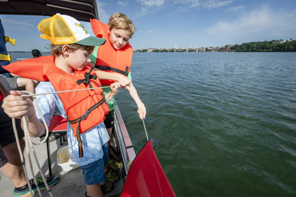 Two boys wearing life jackets and standing on a boat pull on a rope that descends into the lake.