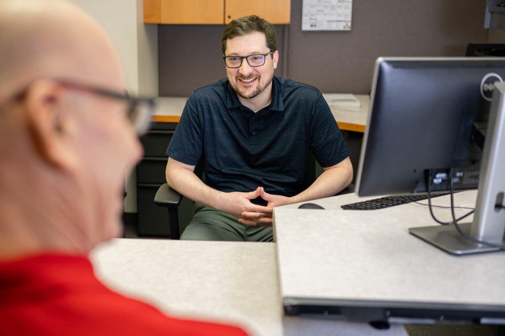 A man sits at a desk and speaks to a person in the foreground. He's smiling.