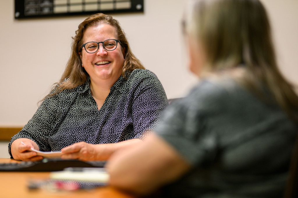 A woman sits in a chair at a table and speaks to someone.