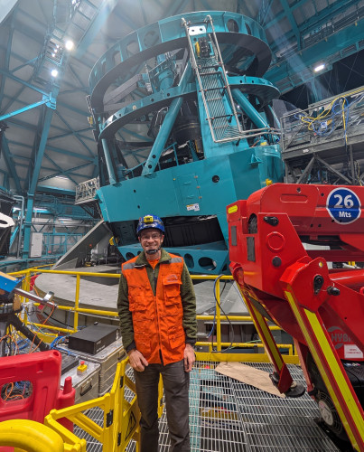 Bechtol wears a bright orange vest, a blue protective helmet and a smile as he stands for a photo in front of the telescope inside the observatory.