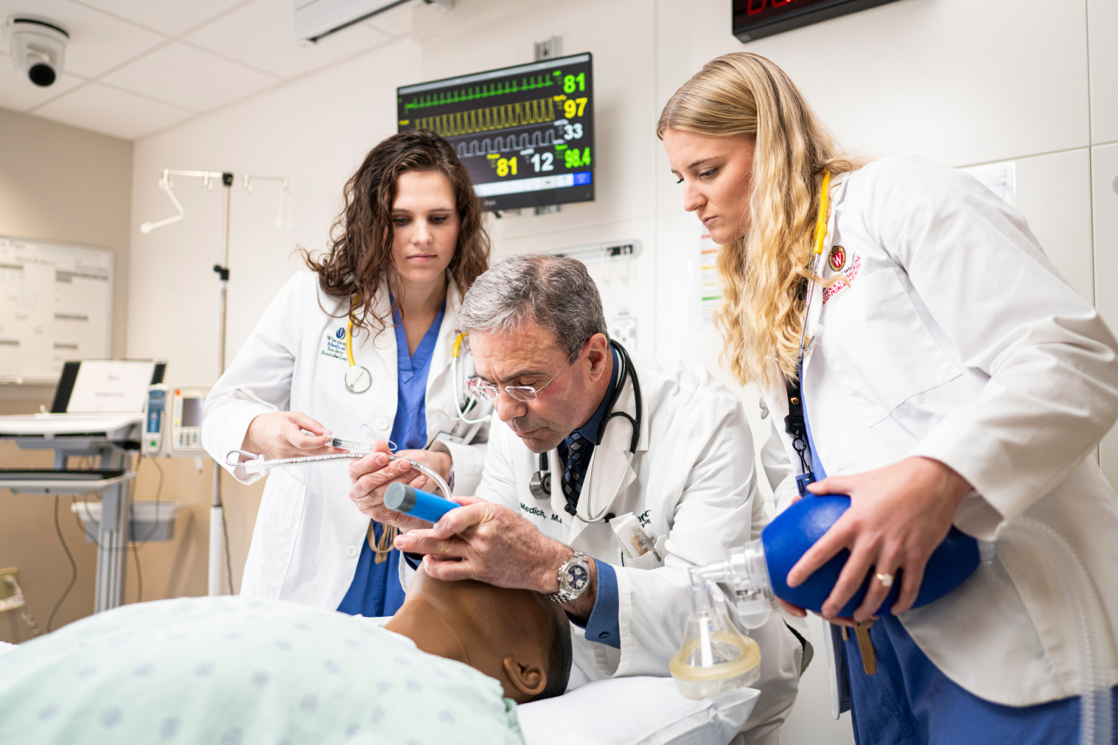 Medich sits on a chair and holds a flexible plastic tube in one had as he leans over a patient that is lying in a hospital bed. Two medical students stand on either side of Medich as he performs the procedure.