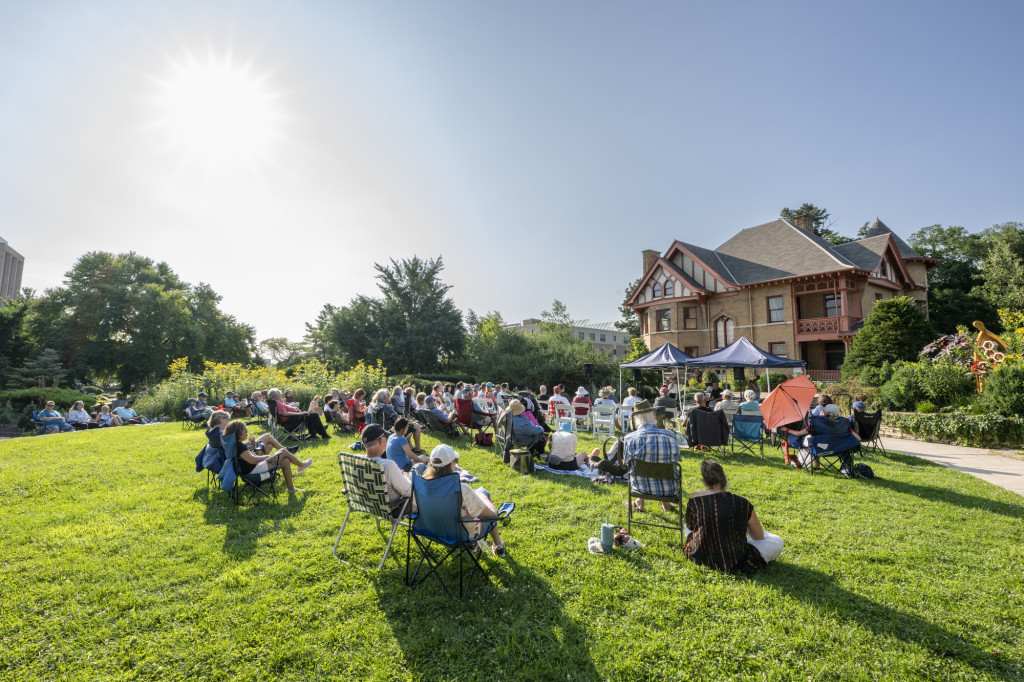 An audience sitting on a lawn listens to a musical group in an outdoor garden setting.