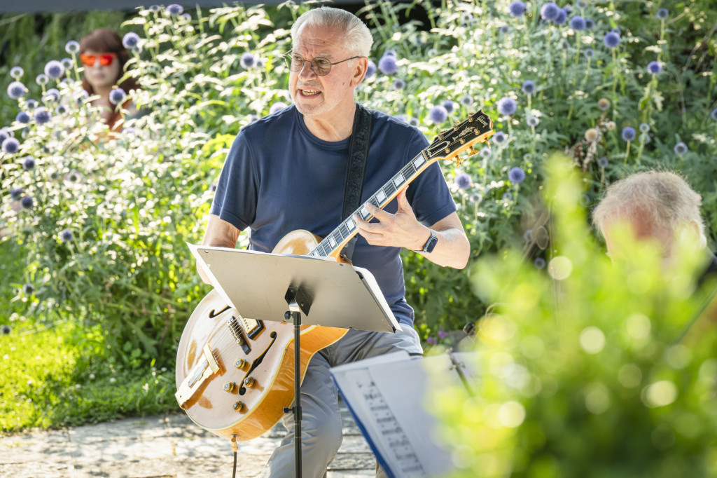 A man strums on a guitar as flowers are visible in the background.f