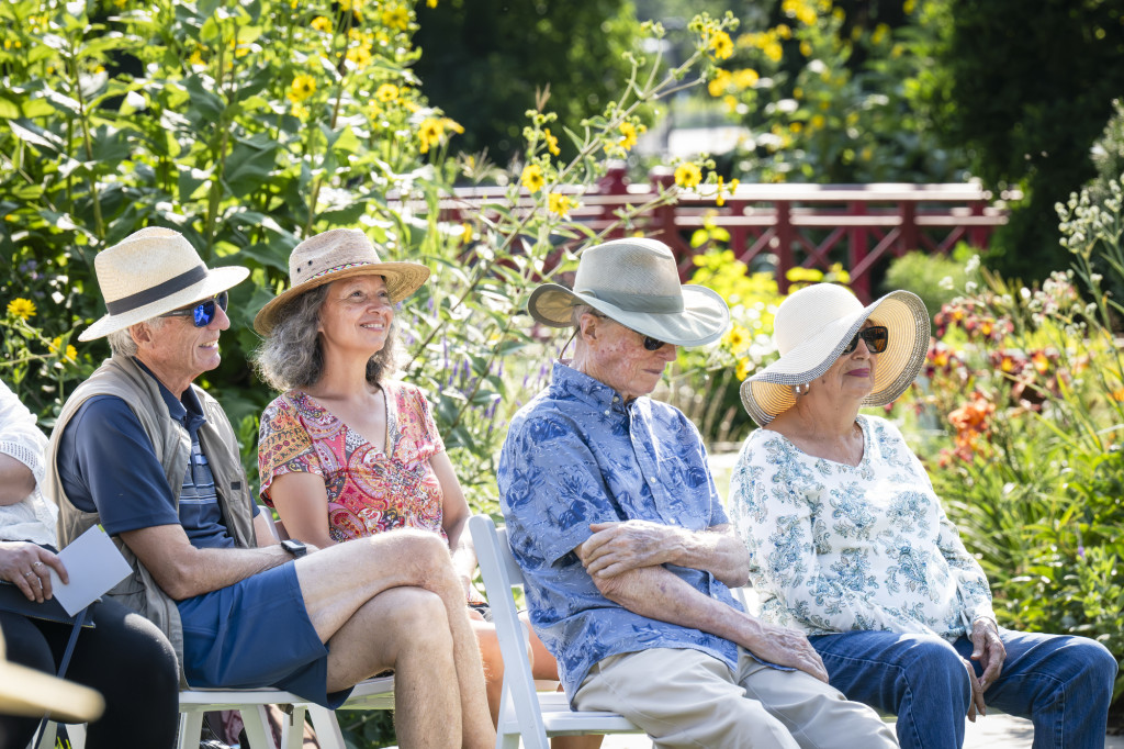 A group of people sit in chairs in the bright sunlight in a garden, listening to a concert.