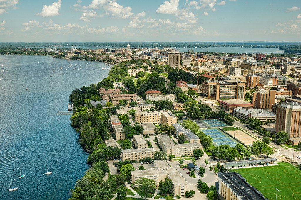 An aerial photograph taken by drone shows the Cole Beach and Lakeshore Tennis Courts and the Lakeshore neighborhood with the Capitol and the campus skyline in the horizon on a partly cloudy summer day. 