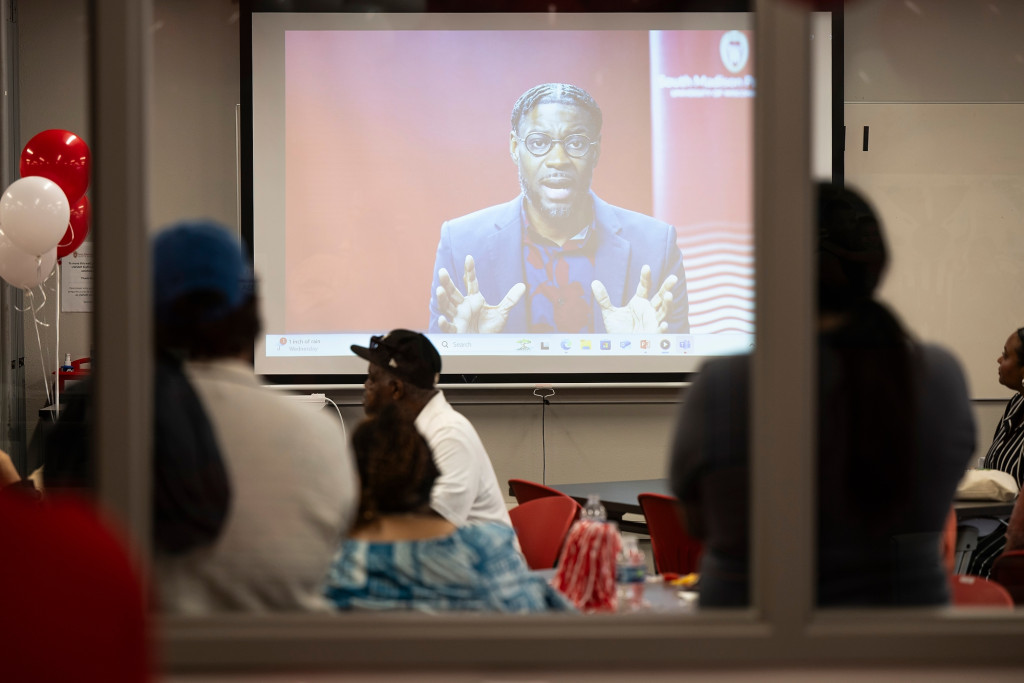 From behind, the photo shows attendees watching a large screen that is projecting a video message from Mitchell.