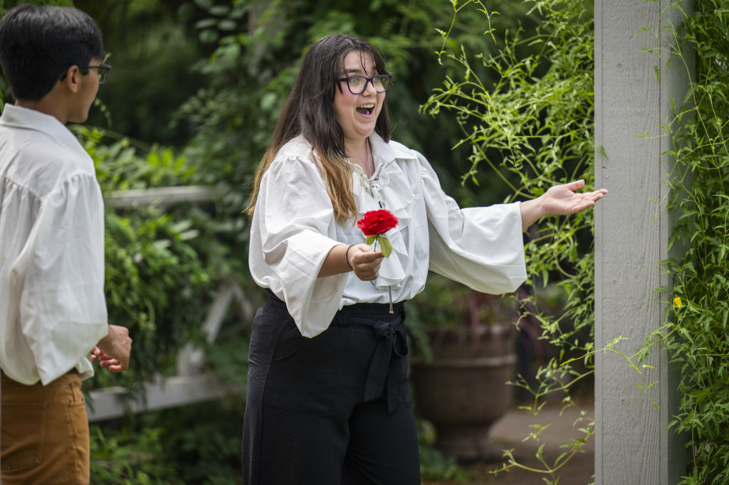 A woman holds a red rose and wears a white shirt as she talks and gesticulates.