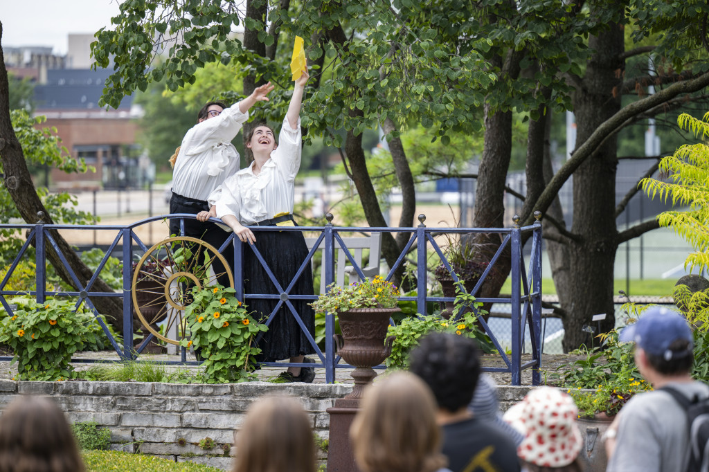 Two people in white shirts act out a scene in a beautiful garden, with an audience watching.