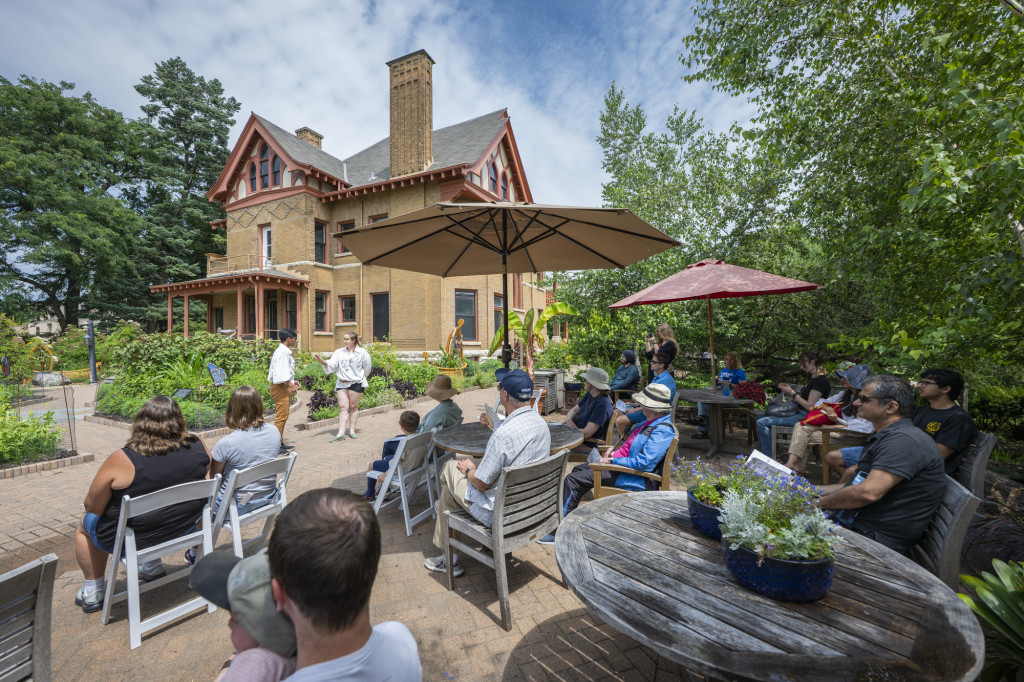 An audience watches a play in a garden with a brick structure in the background.
