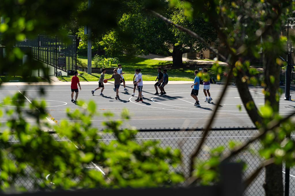 A group of people play basketball on a court surrounded by grass and trees,