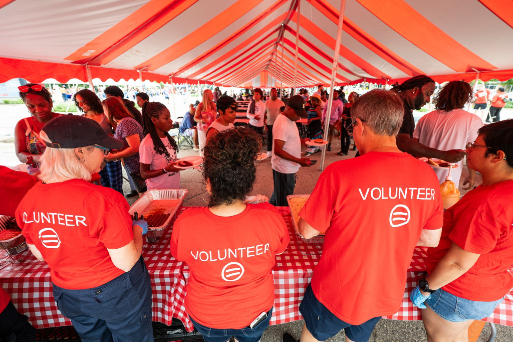 A photo taken from behind shows a group of volunteers all wearing the same shirt and standing in a line in front of a table where food is being served.