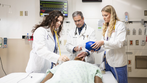 Doctor Michael Medich demonstrates procedure on medical dummy lying on a gurney to two medical students, all three wearing white lab coats