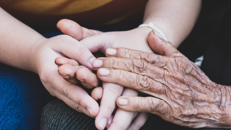 A close up photo shows two people holding hands. One pair appears to be young and the other appears to be older with wrinkles.