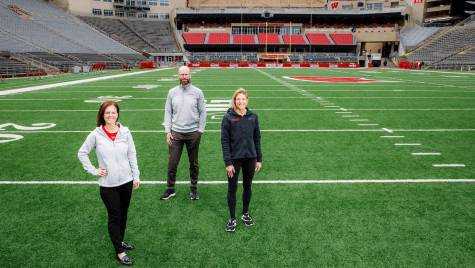 Maria Dehnert, Peter Miller, and Sara Jimenez Soffa stand at the 20 yard line in an empty Camp Randall stadium.