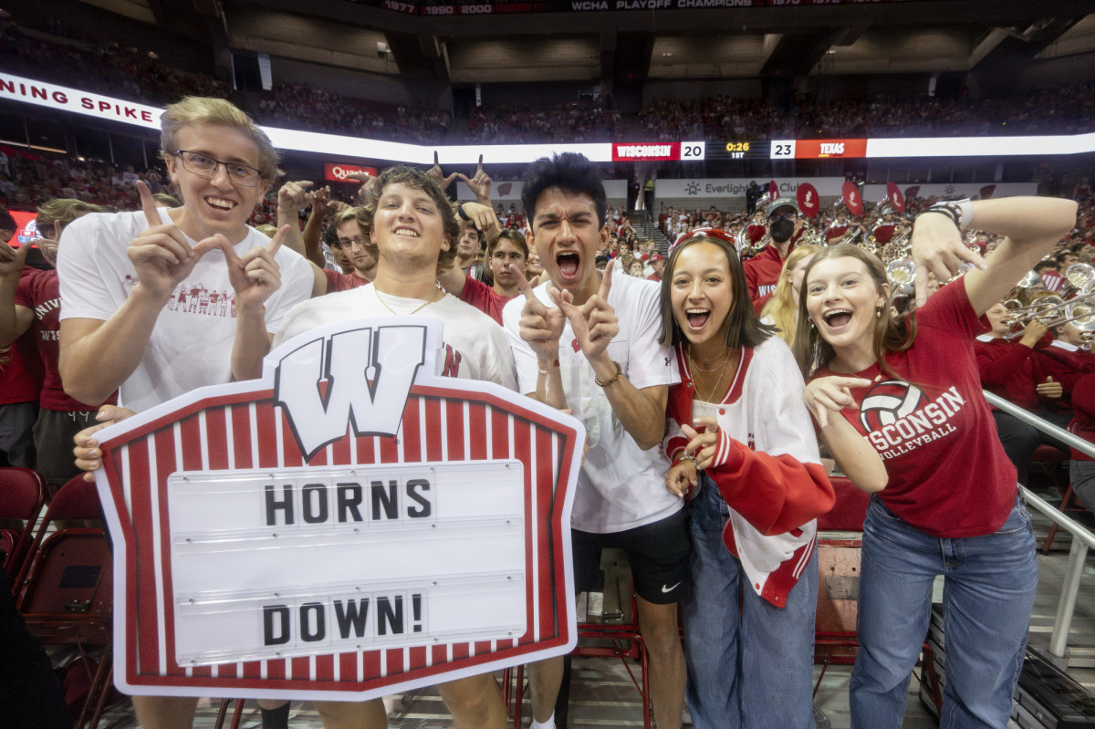 Five Badger fans cheer for the Badgers, making 'w' signs with their hands. One fan holds up a sign in support of the Badger team.
