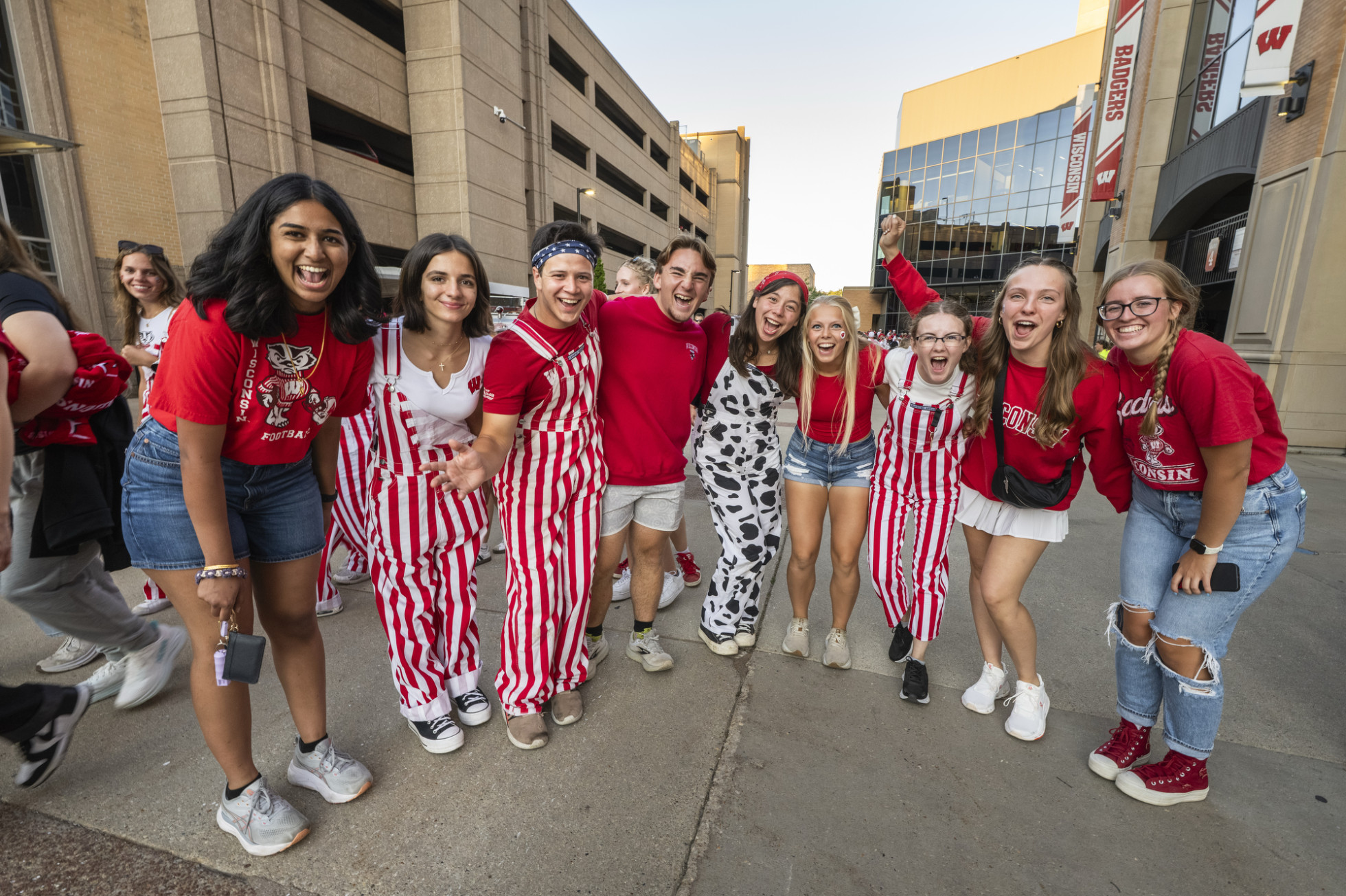 Students wearing Badger gear and red and white overalls pose arm-in-arm outside of Camp Randall Stadium. 