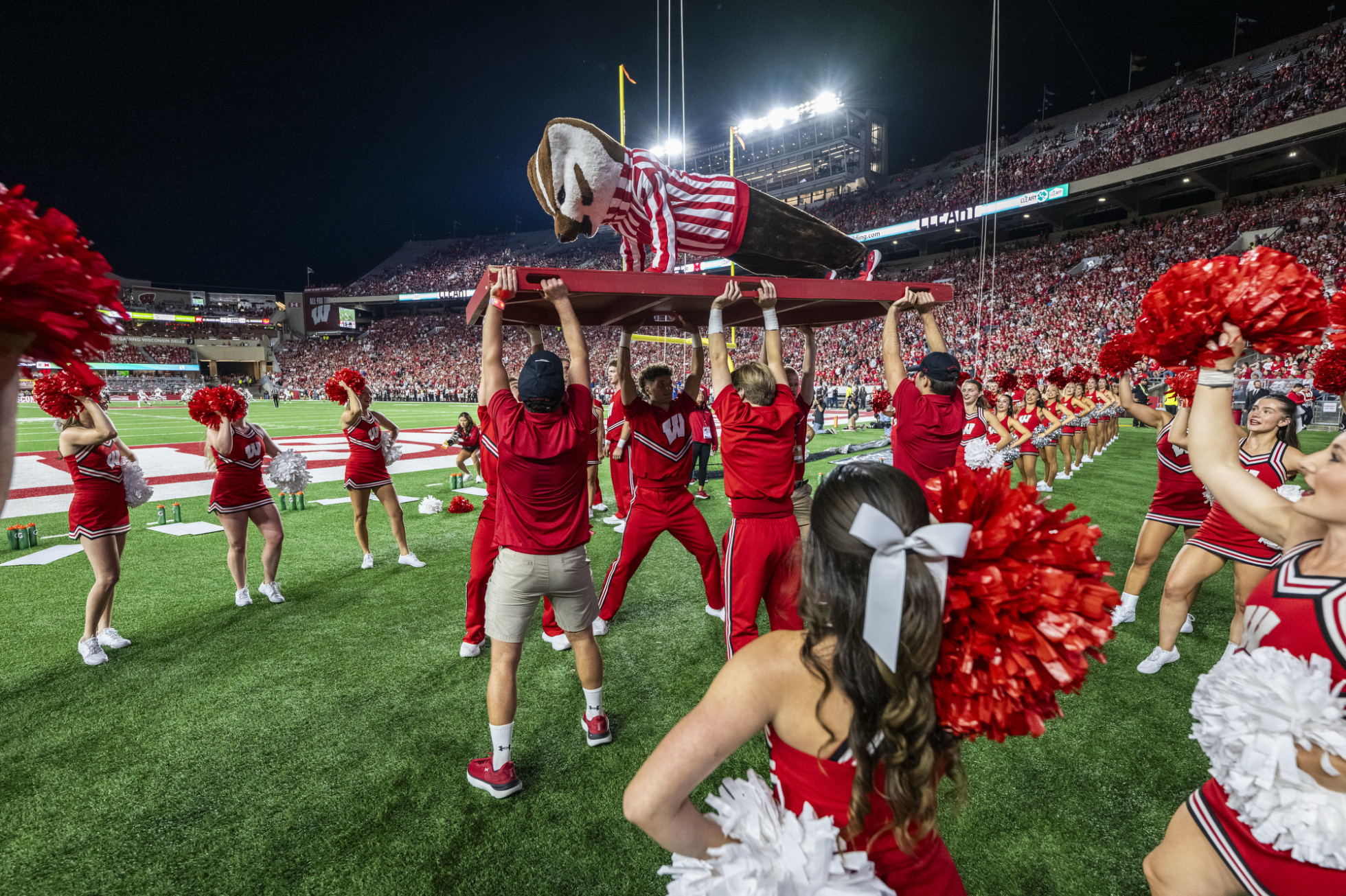Bucky Badger does a push-up on a raised platform on the sidelines, held by UW Spirit Squad members. 