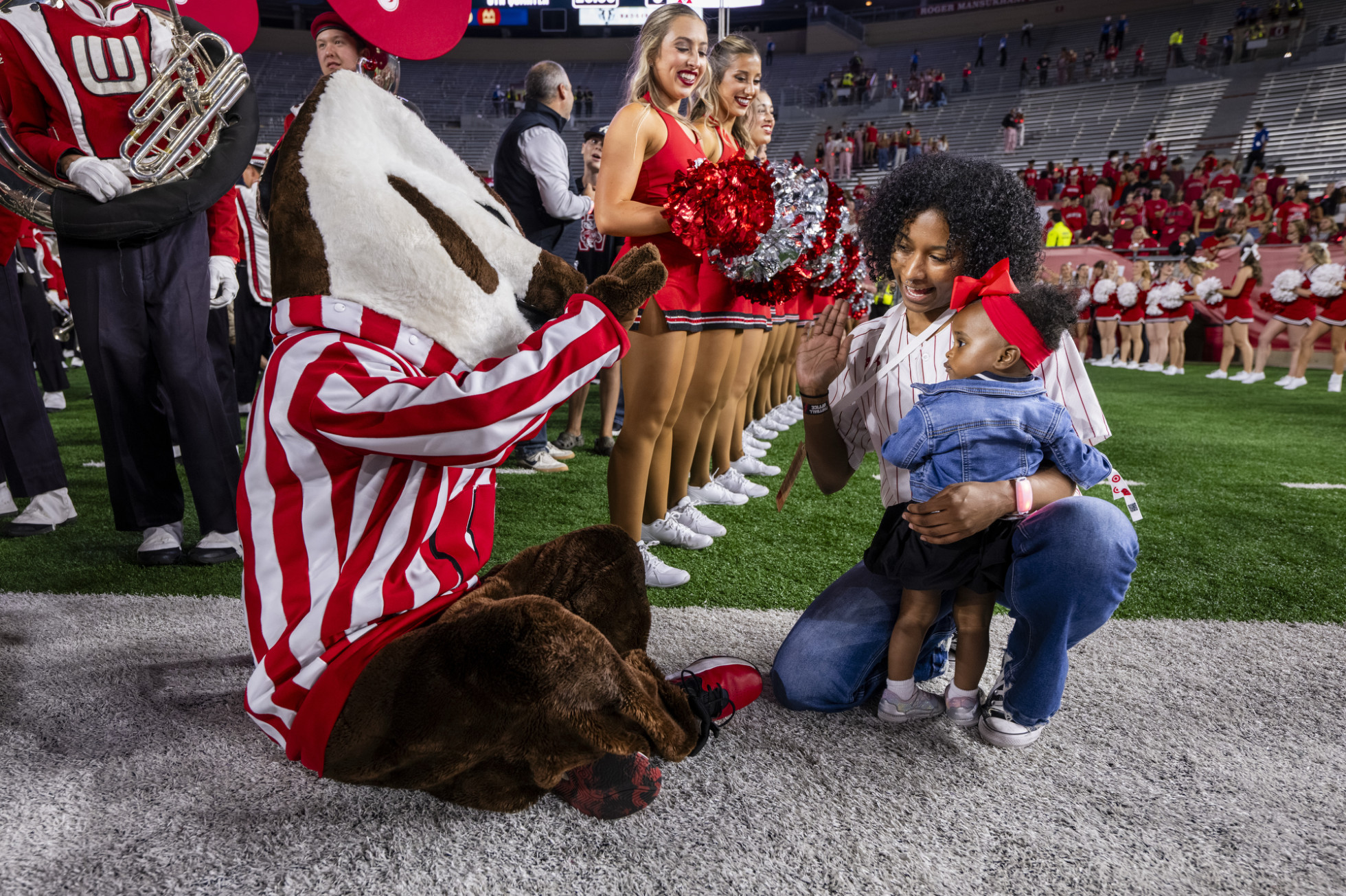 Bucky Badger waves to a a young girl and her mother on the field, next to UW cheerleaders.