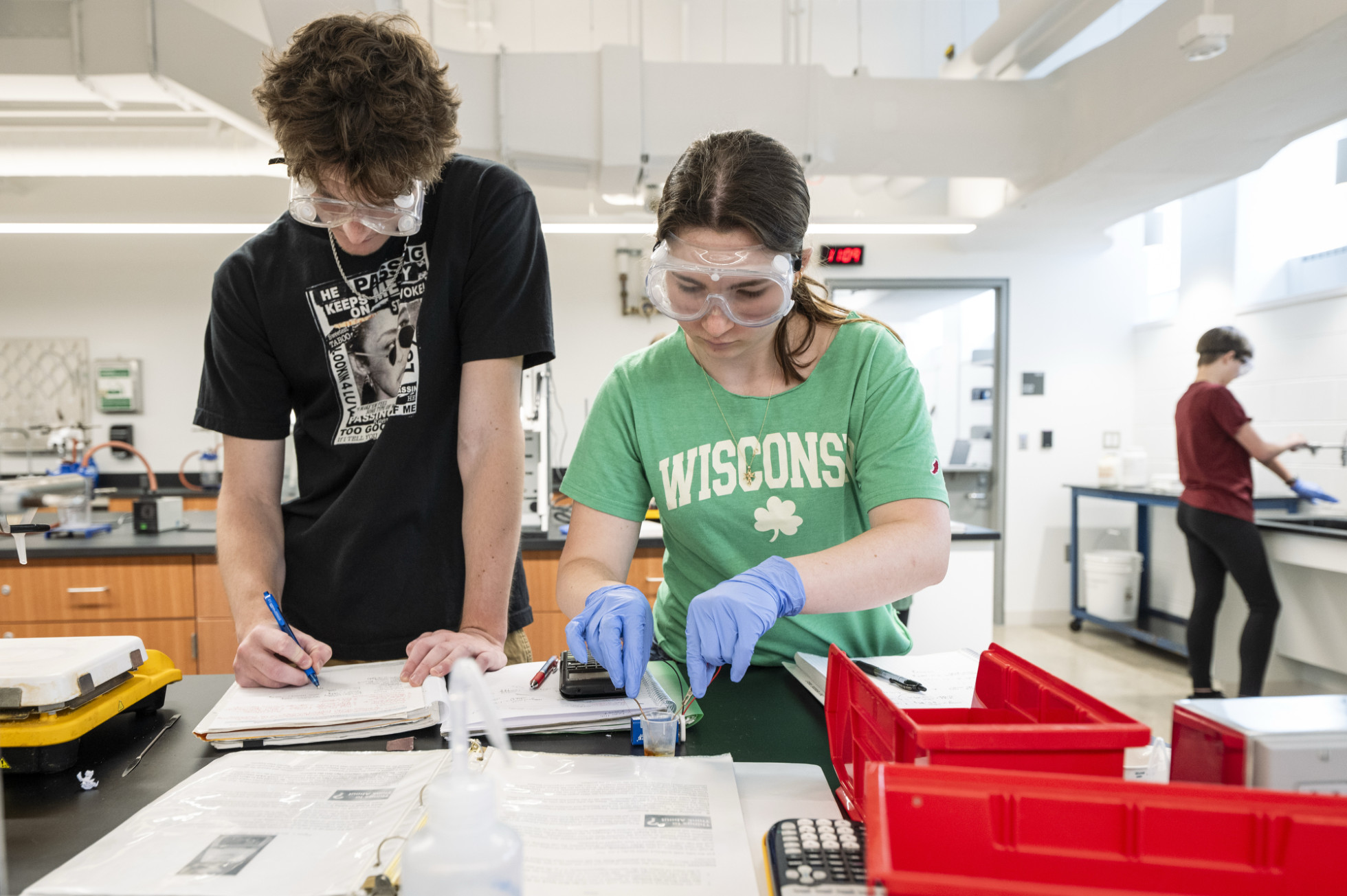 Two students wear safety goggles as they conduct a chemistry experiment. One holds tweezers to connect the 9-volt battery while the other writes observations in a notebook.