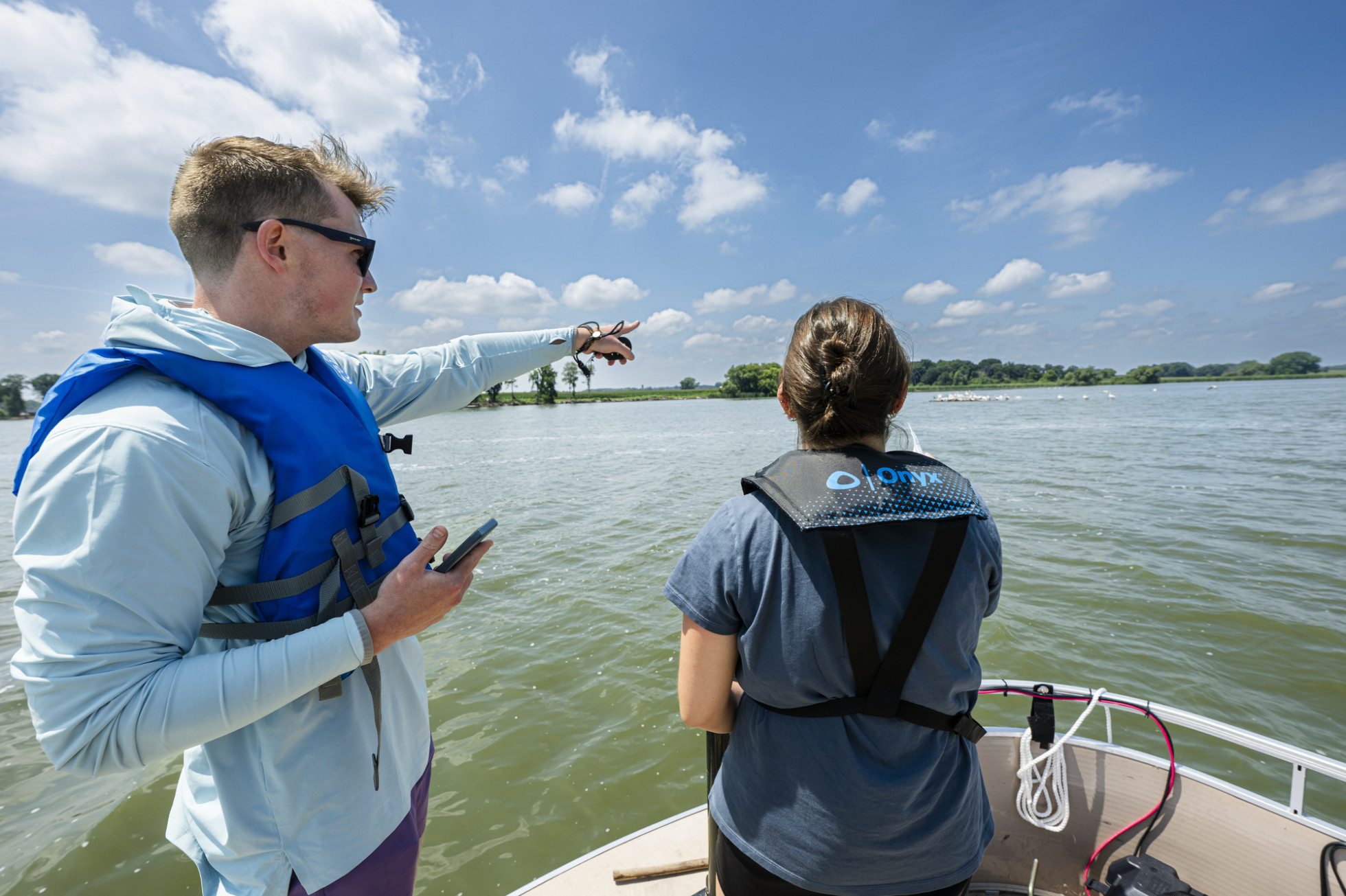 Peterson and Guzman stand on a boat in the middle of a lake. Peterson points to the nearby shoreline.