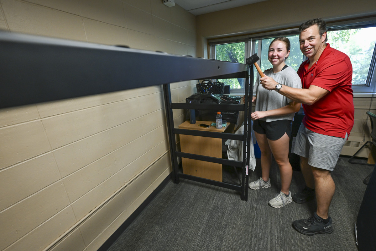 A Smiling Reagan Herdrich and her dad, holding a mallet, assemble a loft bed inside a residence hall room. 