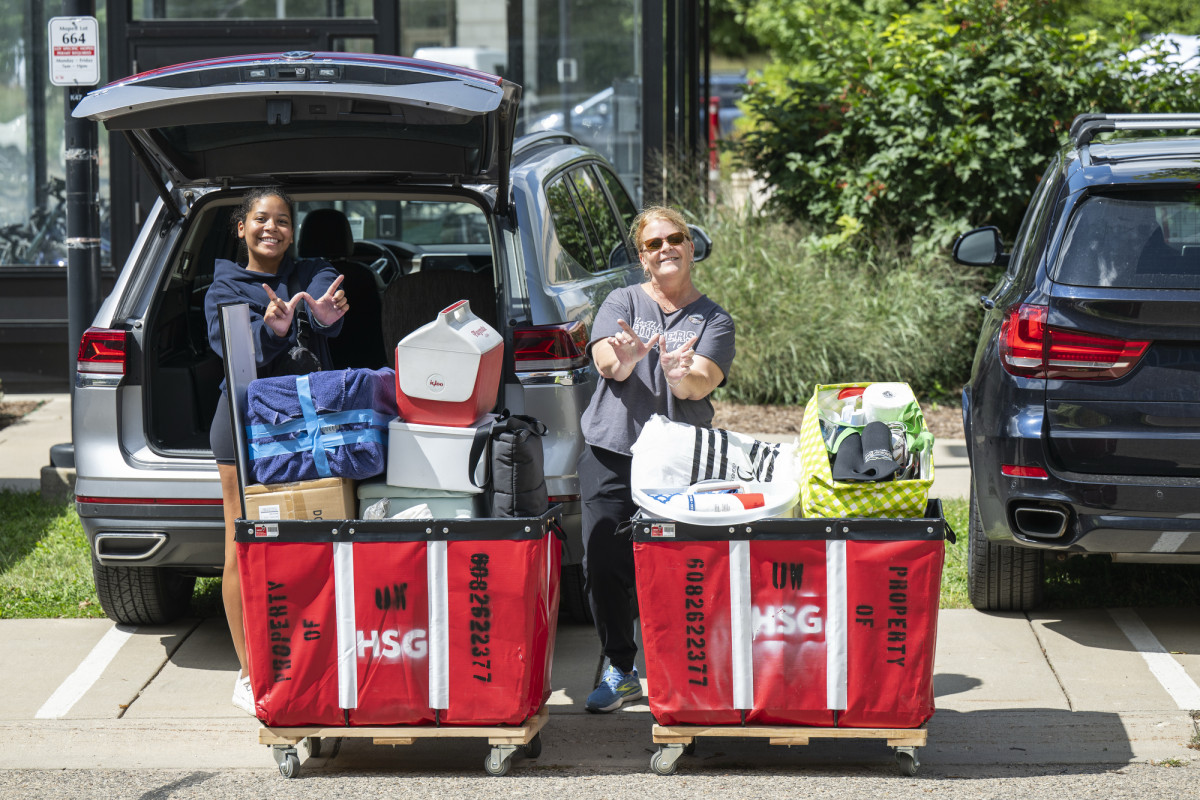 Leena Brown and her mom make Ws with their hands in front of their unpacked car and behind two large red moving cards filled with items. 