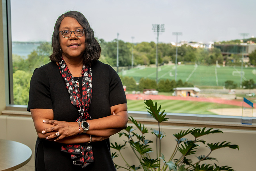 A woman stands in front of a window showing green fields; she's smiling.