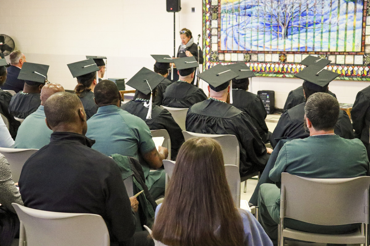 A group of people, some wearing graduation gowns, sit in a classroom and listen to a speaker. It's a commencement ceremony.