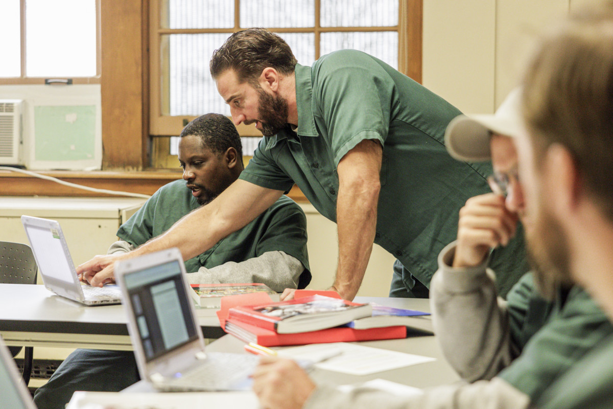 Three men sit at a table and talk and work on a laptop.