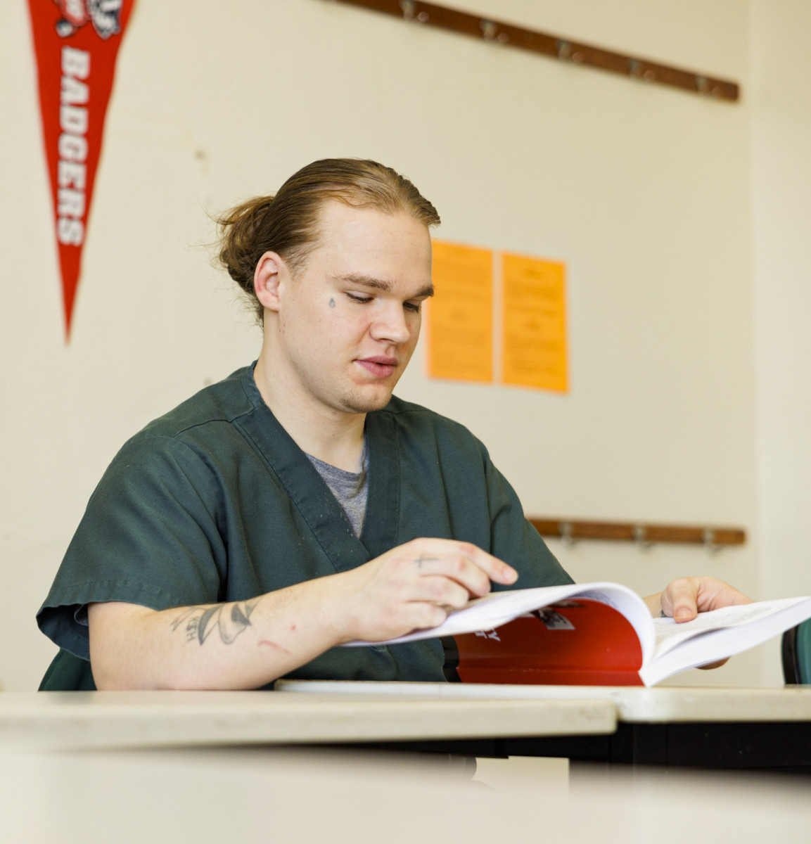 A man wearing a green shirt sits at a table and looks through a book. 