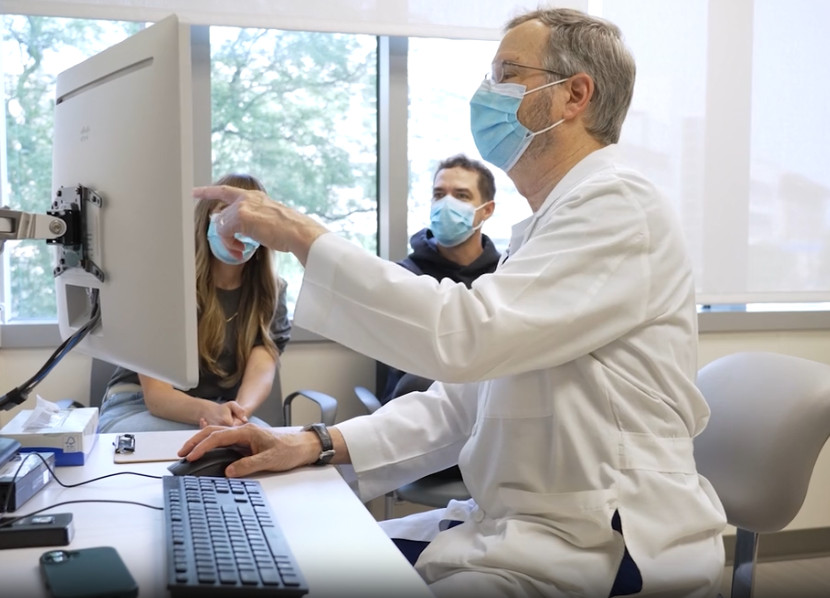 A doctor speaks to two people seated at his desk and points out information on his computer screen. All are wearing masks.