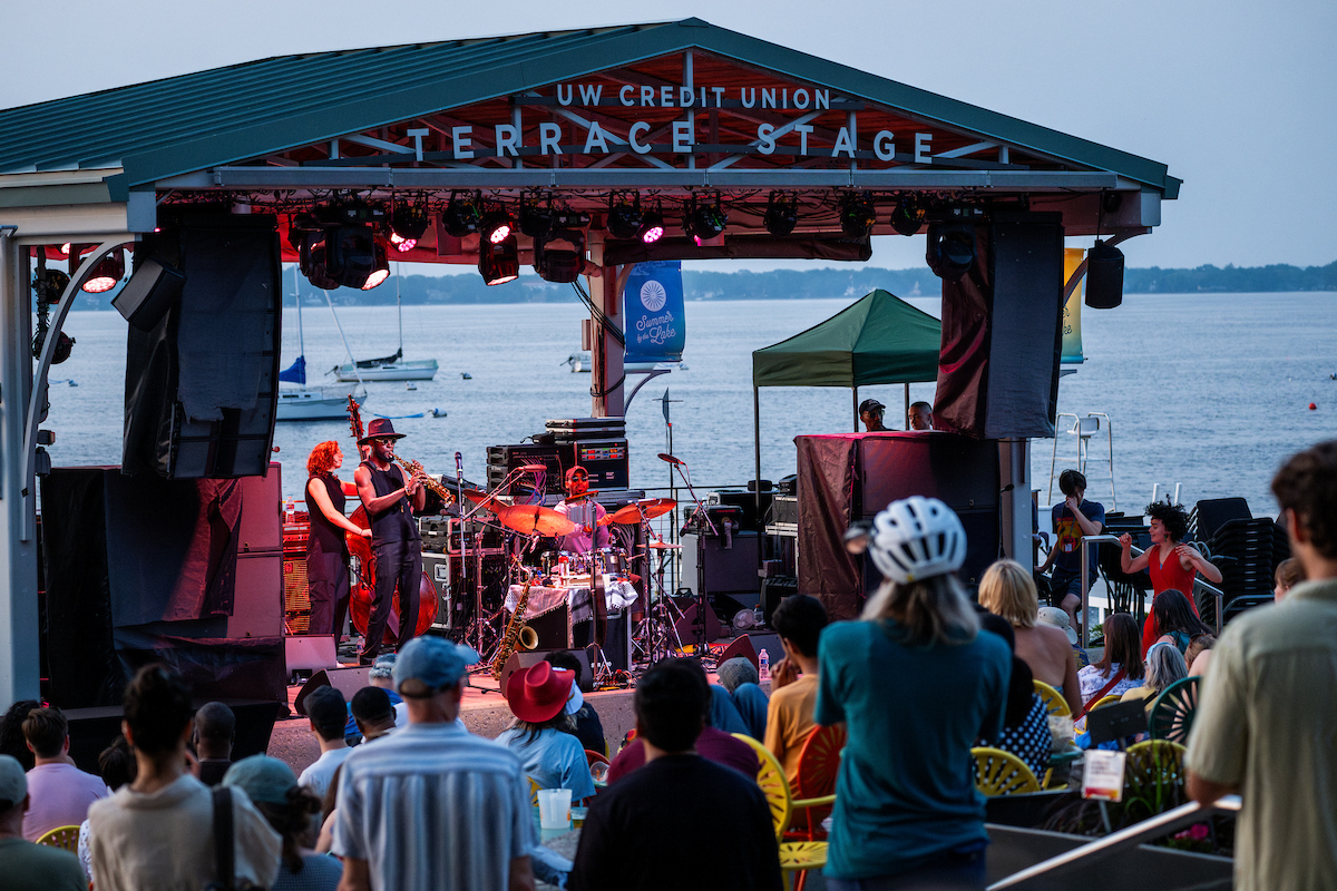 A band plays in a bandshell with a lake visible in the background and people enjoying the music in the foreground.
