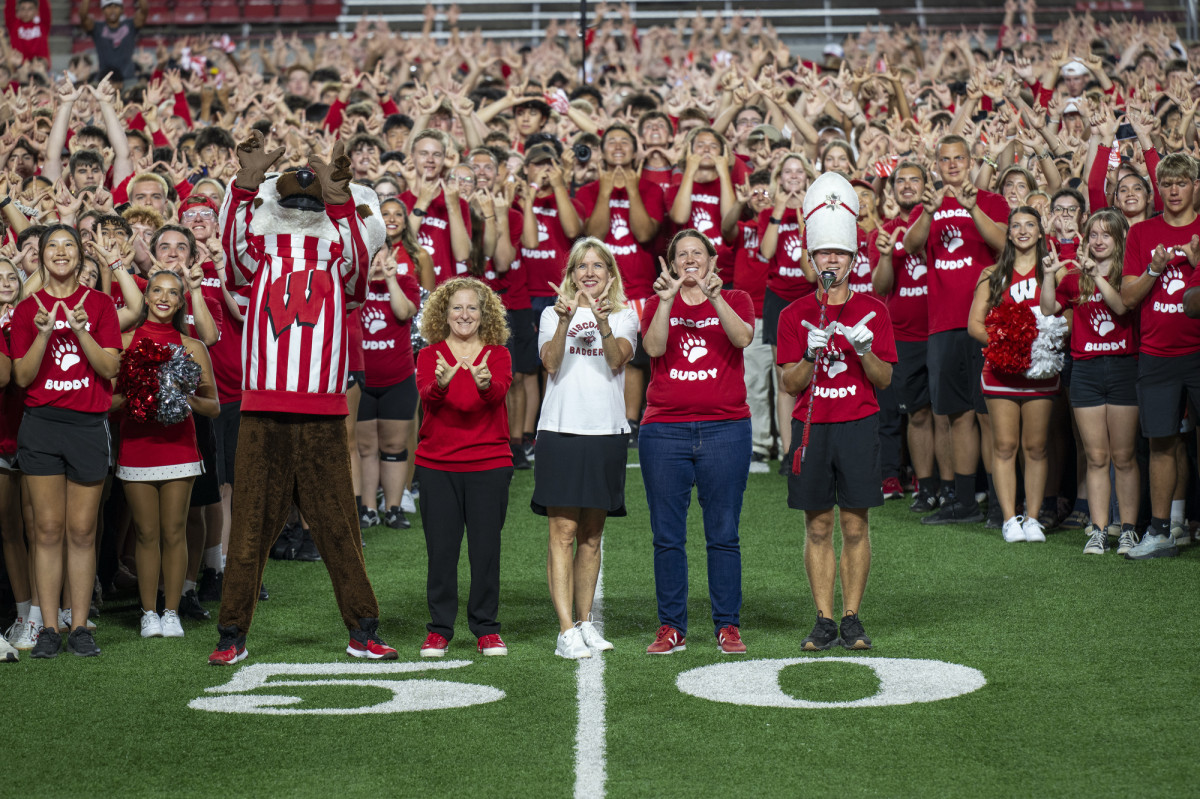 A number of people stand in front of a group of students forming a W.