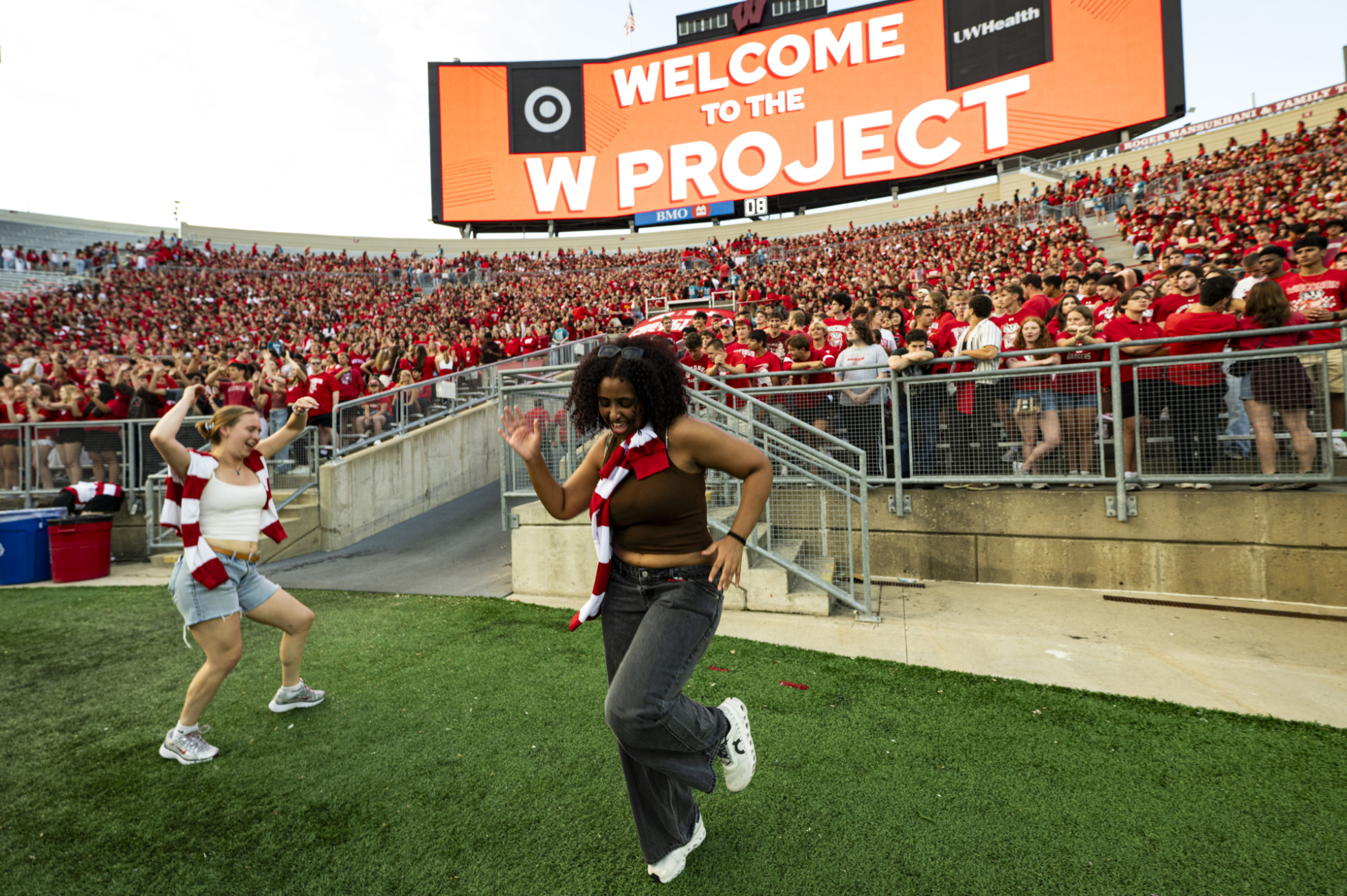 Two students dance on a football field as others watch from the stands.