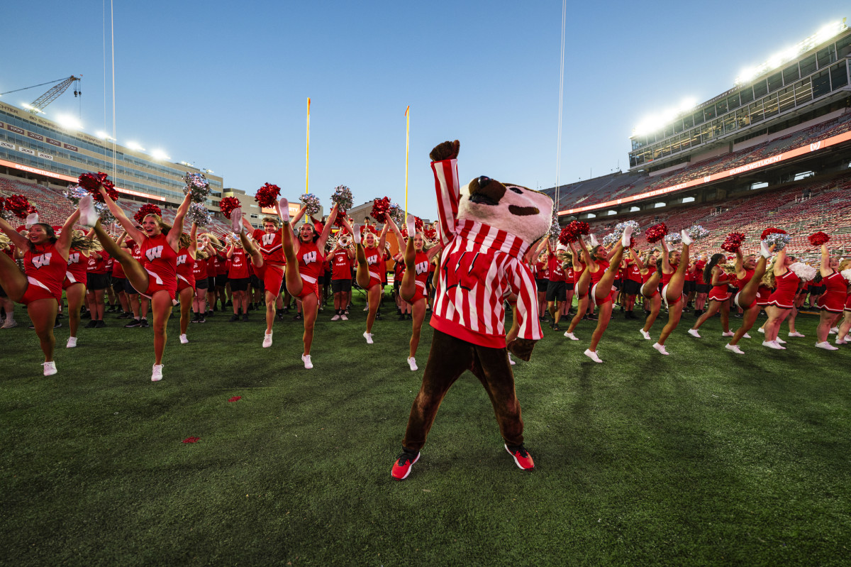 The mascot Bucky Badger holds up his fist and cheers as he's surrounded by cheerleader.