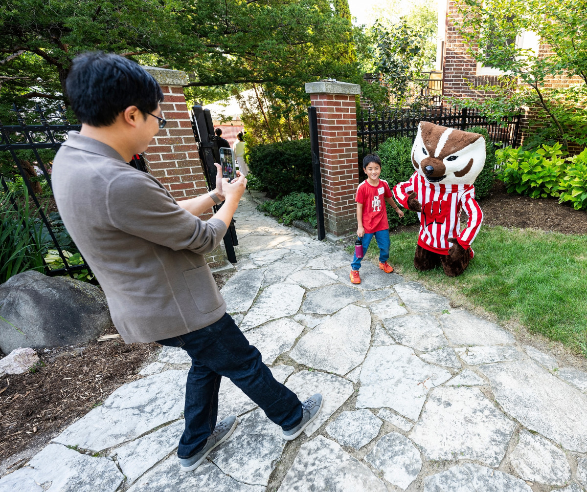 A man stands with a phone in his hands. He aims the phone at his son who is standing next to a kneeling Bucky Badger mascot at the entrance of Olin House.