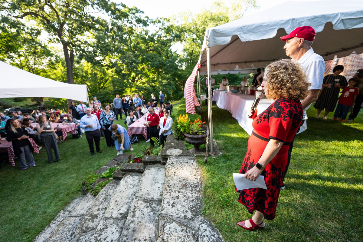 Mnookin, standing next to Foa Dienstag, holds a microphone in her hand and address the crowd of new faculty standing among picnic tables in her backyard.