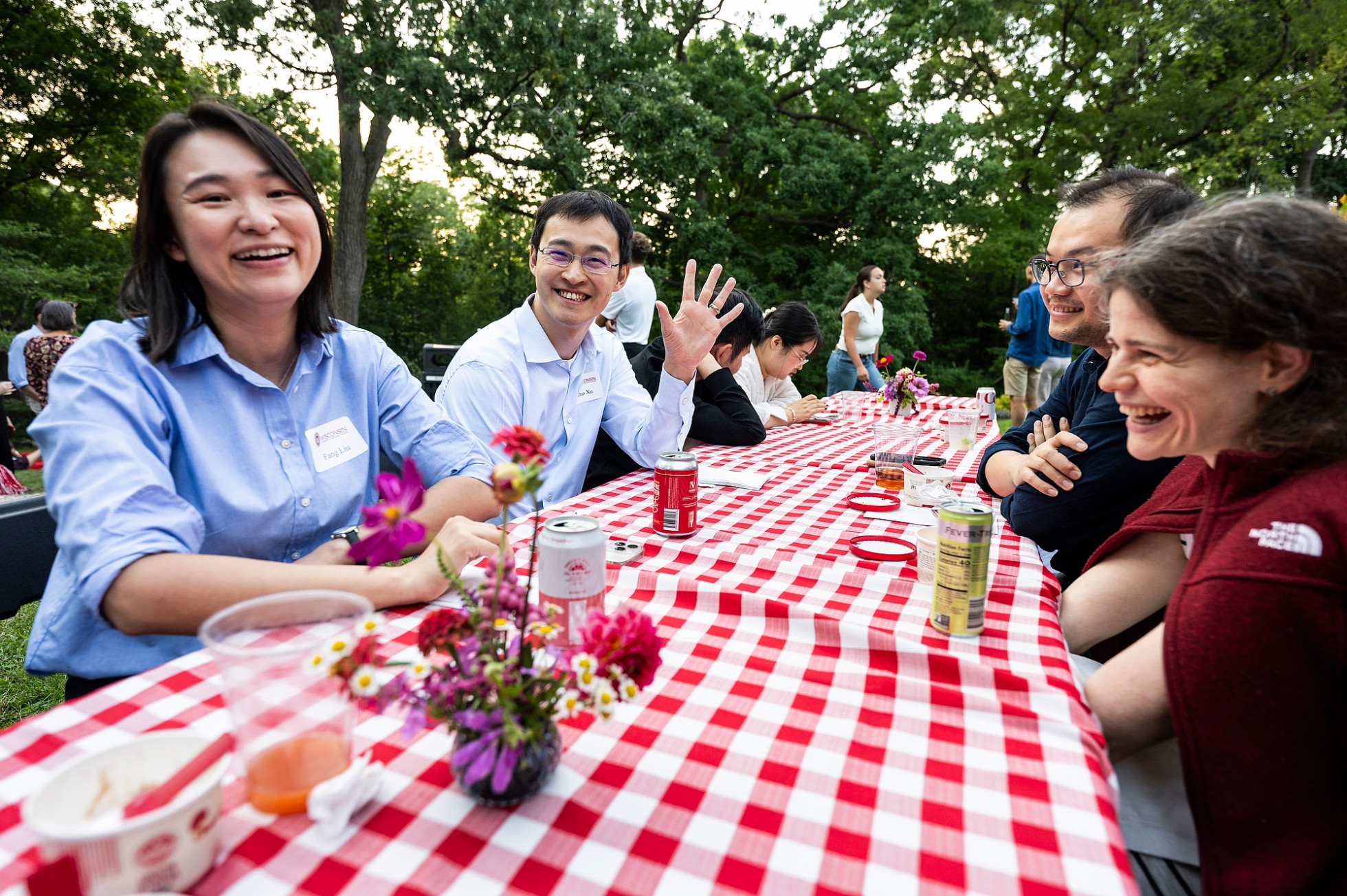 A group of new faculty sitting at a picnic table smile and wave at the camera.