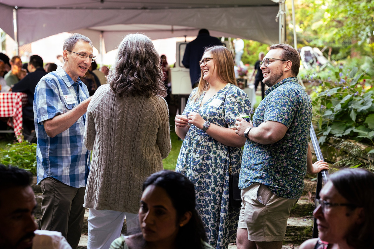 A group of four new faculty smile and laugh while standing in conversation with one another.