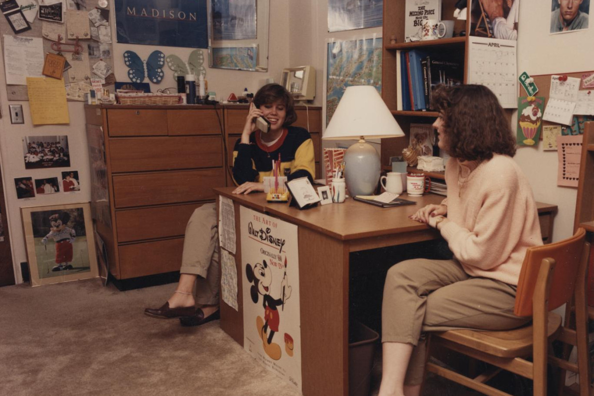 Two students are shown sitting at opposite sides of table, one speaking on a telephone, in a room decorated with posters.