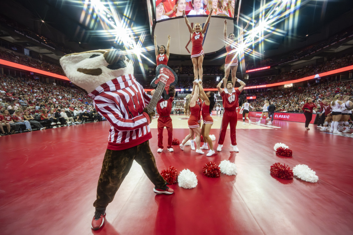Bucky Badger holds up a guitar prop on the volleyball court. Behind him, the Wisconsin Badgers Spirit Squad stand in their cheer formations.
