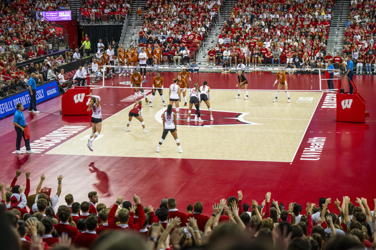 A zoomed out photo shows the Badger women's volleyball team and the Texas Longhorns women's volleyball team on a volleyball court. The court is surrounded by cheering fans.