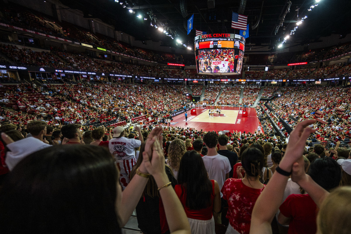 A zoomed out photo shows the Badger women's volleyball team and the Texas Longhorns women's volleyball team on a volleyball court. The court is surrounded by cheering fans.