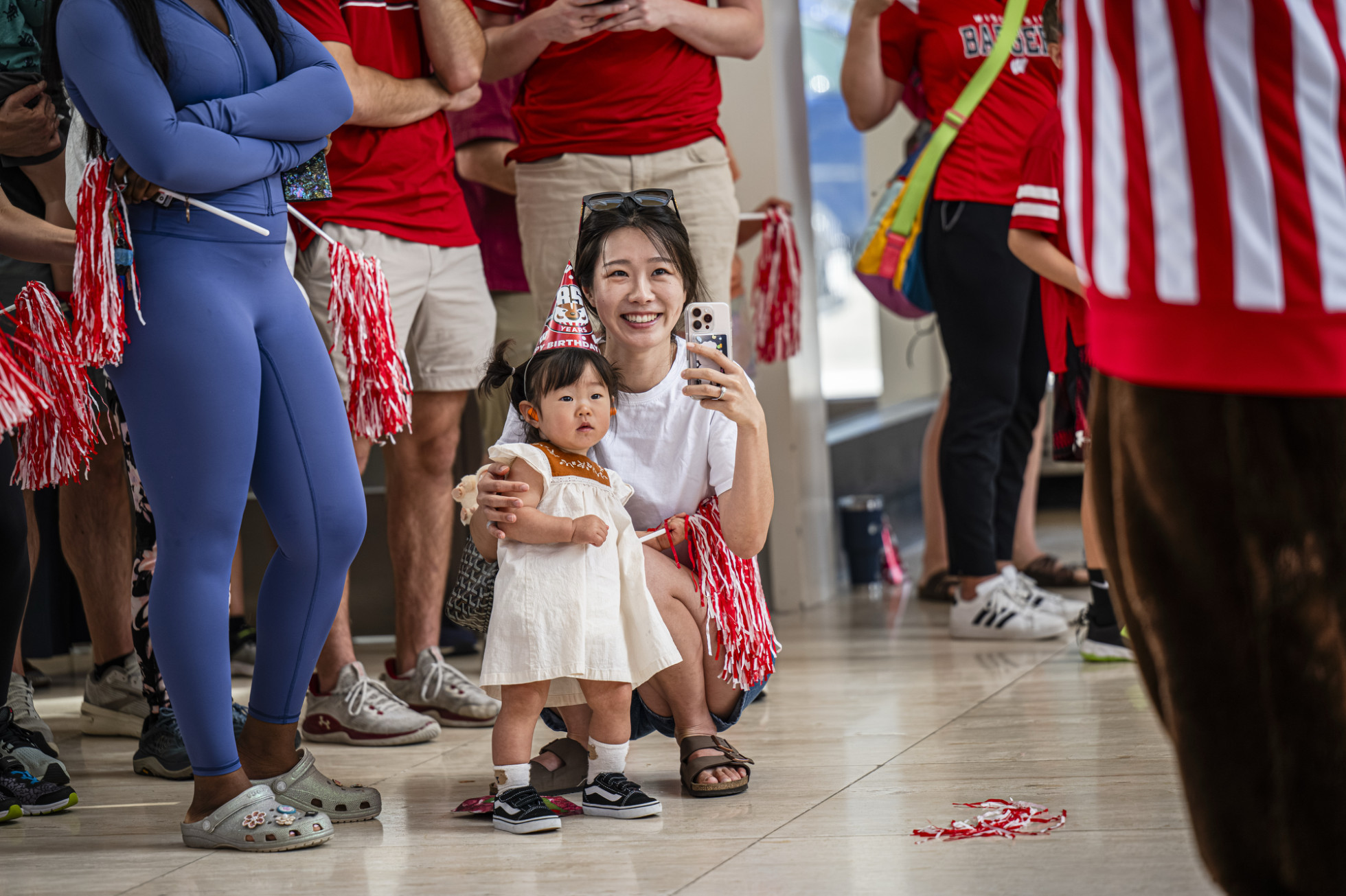 A woman kneels next to her child to watch Bucky Badger perform, holding a phone out to record the action.